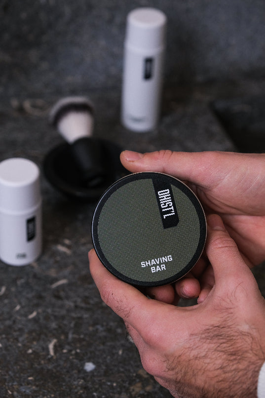 Hand holding a black shaving bar container with 'DUST!' branding on a dark surface.